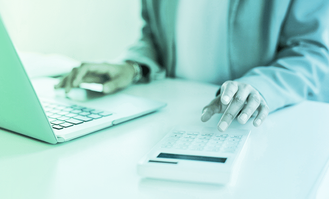 close up picture of a man at his desk, working on a laptpop and a calculator.